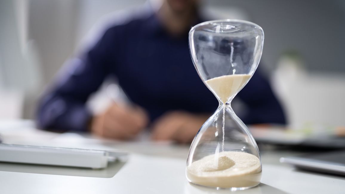 Im Vordergrund rinnt Sand durch eine Sanduhr. Im Hintergrund sitzt eine verschwommen zu sehende Person vor einem PC und notiert etwas.