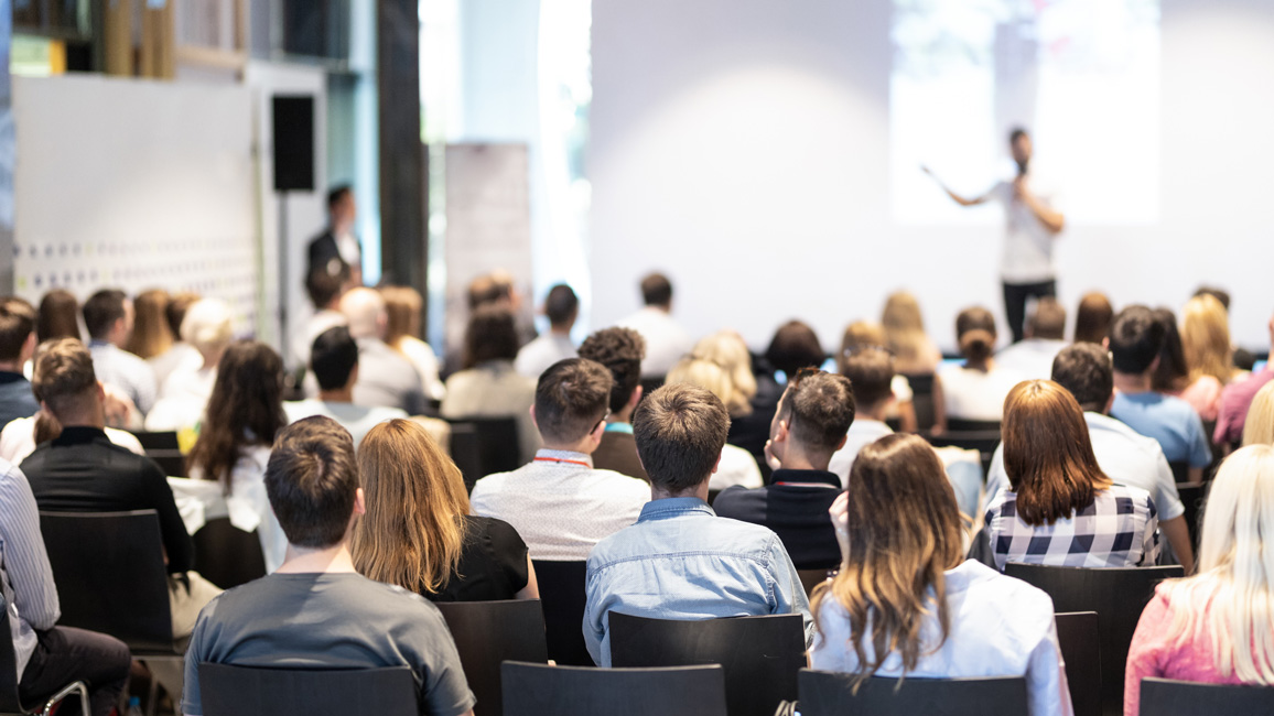 Blick aus den Reihen der Teilnehmenden einer Konferenz auf einen verschwommen zu sehenden Sprecher im Hintergrund.