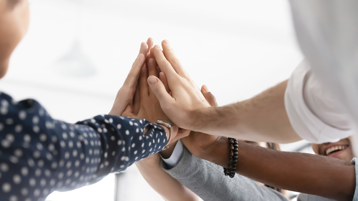 Several men and women of different ethnological backgrounds put their hands on top of each other, forming a small cone.