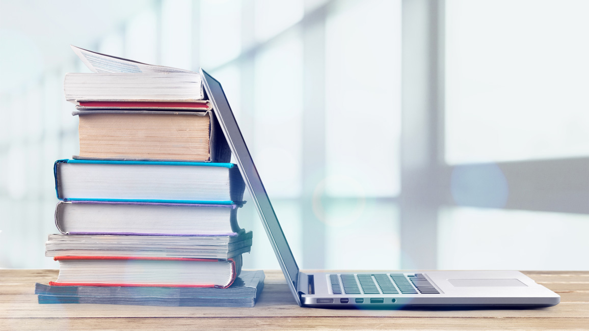 A laptop on a table. Its open monitor is leaning against a pile of books.