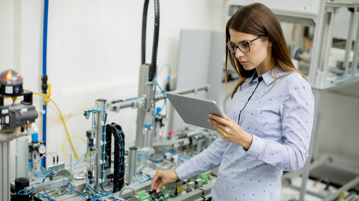 A young woman holding a tablet is standing next to a complex apparatus, checking its settings.